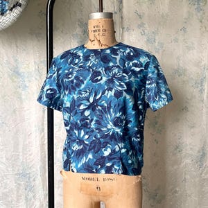 May include: A short-sleeved top with a floral pattern in shades of blue and white. The top is displayed on a vintage dress form. The floral design features large, stylized flowers.