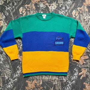 May include: A color-blocked sweater in green, blue, and yellow. The sweater features a crew neck and long sleeves with a ribbed cuff. A small patch with the Lacoste logo is on the chest. The sweater is made of a soft knit material.
