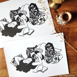 May include: Two black and white art prints depict a person reclining and reading, surrounded by pillows with floral patterns. The prints are on white paper, with a wooden table in the background. The text "Atelier de l'Hermine - Avril 2025" is visible.