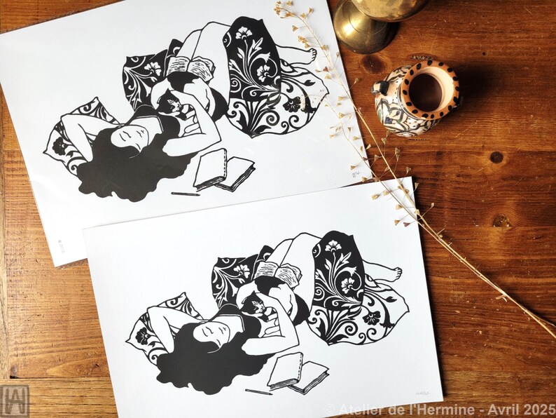 May include: Two black and white art prints depict a person reclining and reading, surrounded by pillows with floral patterns. The prints are on white paper, with a wooden table in the background. The text "Atelier de l'Hermine - Avril 2025" is visible.