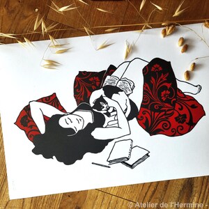 May include: Black and white art print depicting a person reclining with a cat, reading a book. The person is surrounded by a red patterned blanket and pillows. Two notebooks and a pencil are in the foreground.