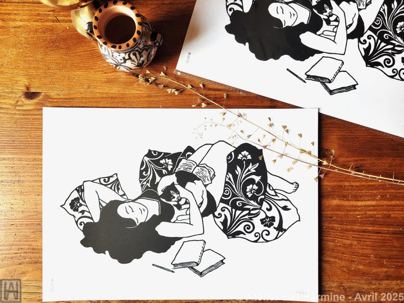 May include: Black and white art print featuring a woman reclining with a book and a cat. The print has a floral pattern on the blanket and pillow. A small ceramic vase and sprigs of dried flowers are on the wooden surface.
