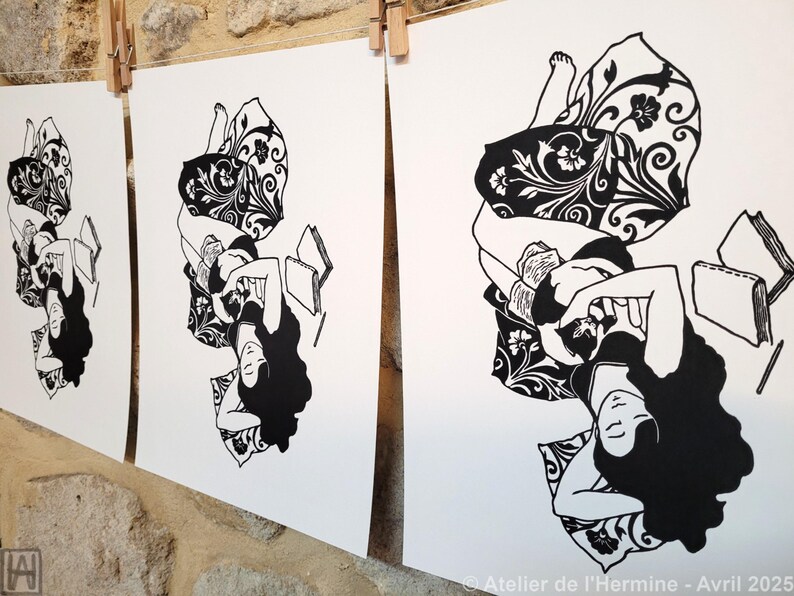 May include: Black and white art prints of a person reclining with a book, featuring long, flowing hair and patterned pillows. The prints are displayed on a clothesline against a stone wall. The artist's signature is visible.