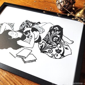 May include: Black and white art print in a black frame. The illustration depicts a person lying down with a cat, surrounded by pillows and a patterned blanket. Two books and a pencil are in the foreground. The artwork is signed.