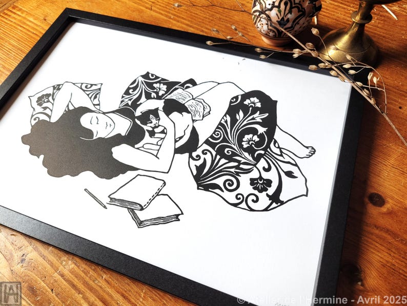 May include: Black and white art print in a black frame. The illustration depicts a person lying down with a cat, surrounded by pillows and a patterned blanket. Two books and a pencil are in the foreground. The artwork is signed.