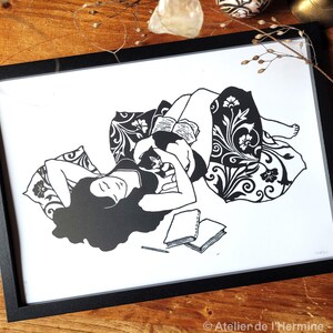 May include: Black and white framed art print depicting a person reclining with a cat, reading a book. The illustration features floral patterns and a relaxed, cozy scene. The print includes the text "Atelier de l'Hermine - Avril 2025".