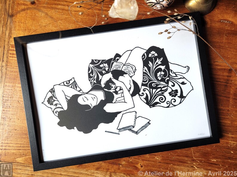 May include: Black and white framed art print depicting a person reclining with a cat, reading a book. The illustration features floral patterns and a relaxed, cozy scene. The print includes the text "Atelier de l'Hermine - Avril 2025".