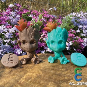 May include: Two Groot-shaped planters, one brown and one teal, each holding faux succulents. The planters sit on a wooden surface with a floral background. Each planter has a matching round base with the text "@CC_CRESTIVE".
