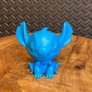 Puede incluir: Figura azul brillante del personaje Stitch de la película animada Lilo & Stitch. La figura está sobre una superficie de madera. El personaje tiene orejas grandes, una nariz ancha y un cuerpo pequeño.