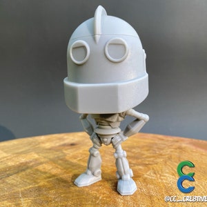 May include: A grey Iron Giant robot figurine, posed with hands on hips. The figure has a rectangular head, round eyes, and a fin on top. The figure is on a wooden surface.