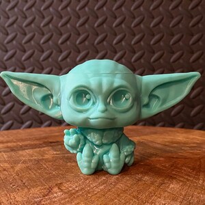 May include: A teal-colored figurine of a character with large ears and eyes, seated with one hand raised. The figure has a detailed face and is made of a solid material. The figurine is a collectible item.
