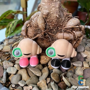 Pode incluir: Duas pequenas figuras impressas em 3D de cor bege com olhos verdes e bocas sorrindo. As figuras têm sapatos rosa e pretos e estão sentadas em uma cama de pequenas pedras cinzas.