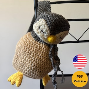 Op de afbeelding: Een handgemaakte gehaakte pinguïntas in grijstinten, beige en geel. De pinguïn heeft gesloten ogen, een gele snavel en gele voeten. De tas heeft een grijze sjaal en een band. De tas hangt aan een zwart metalen rek.