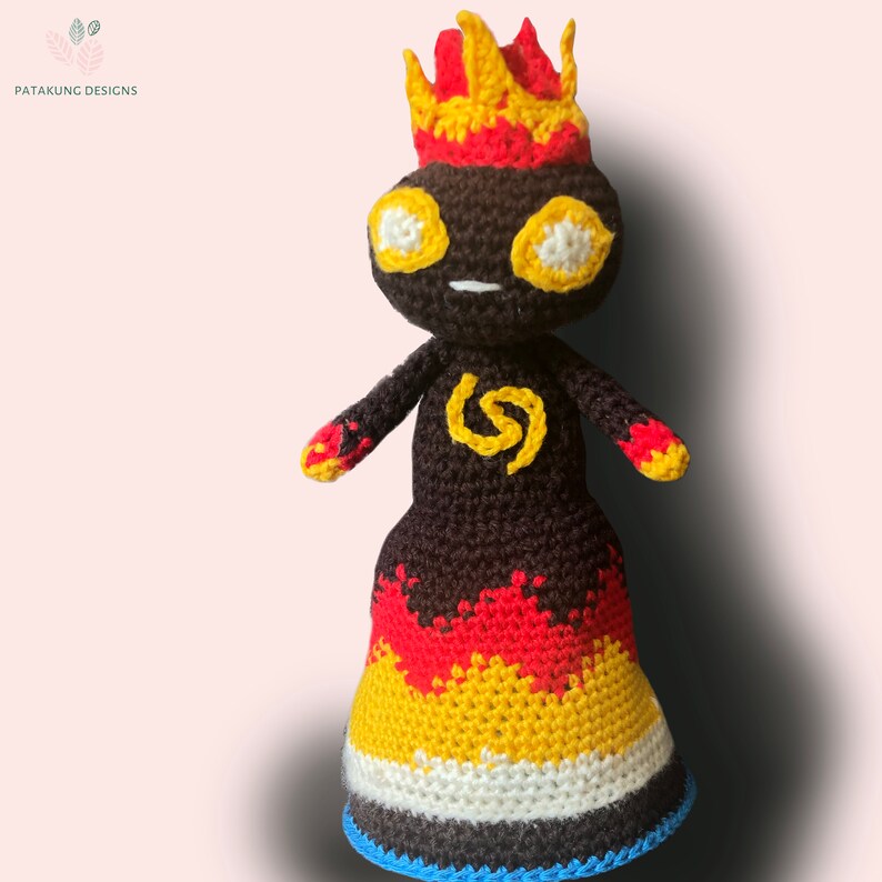 A crochet volcano doll with fiery hair and eyes and a yellow spiral symbol on her chest against a light pink background.
