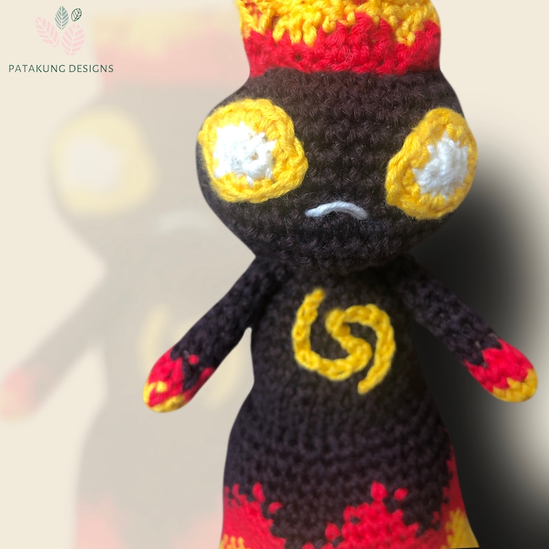 A close-up view of the crochet volcano doll with fiery hair and eyes and a yellow spiral symbol on her chest with a shadow behind the doll.