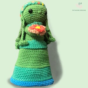 A side view of the crochet green Mother Earth doll holding a pink/red flower with a floral headband against a light green background.
