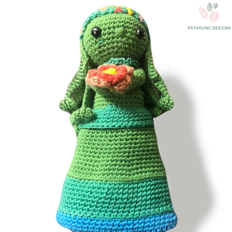A crochet green Mother Earth doll holding a pink/red flower with a floral headband against a white background.