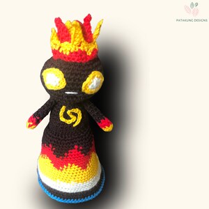 A slight top view of the crochet volcano doll with fiery hair and eyes and a yellow spiral symbol on her chest against a light yellow background.