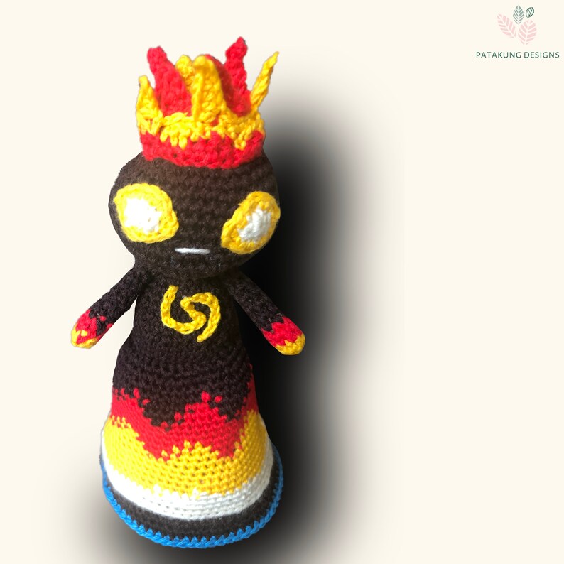 A slight top view of the crochet volcano doll with fiery hair and eyes and a yellow spiral symbol on her chest against a light yellow background.