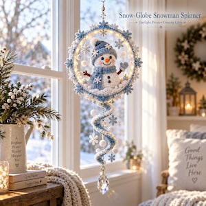 May include: A hanging Snow-Globe Snowman Spinner decoration featuring a crocheted snowman in a blue hat and scarf inside a circular frame adorned with blue and white snowflakes. A spiral tail hangs below with a clear crystal prism at the bottom.
