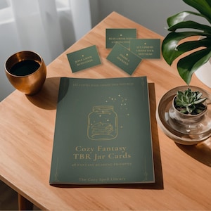 Puede incluir: Un folleto de color verde oscuro titulado Cozy Fantasy TBR Jar Cards con 48 sugerencias de lectura de fantasía, que presenta una ilustración de un frasco dorado y varias tarjetas rectangulares a juego con textos como Judge a Book by Its Cover.