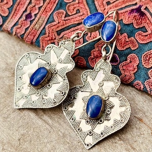 以下が含まれることがあります： ハート型のデザインの銀色のイヤリング。複雑な模様と、中央に楕円形の濃い青色の石が特徴です。追加の小さな青い石がハートの上に配置されています。イヤリングは、背景にカラフルなテキスタイルがある、テクスチャ加工された表面に表示されています。