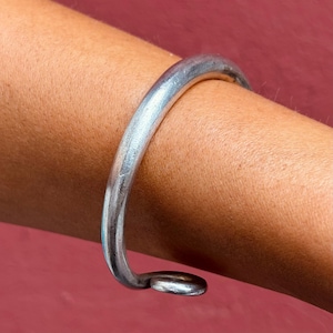 Könnte beinhalten: Ein silberfarbenes, gebogenes Armreif-Armband. Das Armband ist ein massives, abgerundetes Band, das sich um den Arm krümmt, mit einer kleinen, abgeflachten Schlaufe am Ende. Das Metall hat ein leicht strukturiertes Aussehen.