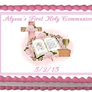 May include: A pink frosted cake with a white and pink border. The cake features a pink rectangular image with the text "Alyssa's First Holy Communion" and the date "5/2/15". The image includes a cross, flowers, a bible, a chalice, and bread.