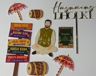May include: A collection of decorative cutouts featuring a man, drums, umbrellas, and signs with text. The signs read "KHAO PIYO", "NACHO GAO", "CHAI SHAI", "KHELO KUDO", and "PHOTO KHEECHO". A gold cake topper says "Hasnains DHOLKI".