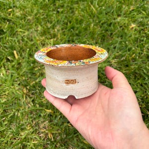 May include: A small, beige ceramic pot with a colorful floral design around the rim. The pot has a cylindrical shape with a slightly flared top and a small rectangular label that reads "mini". The pot is held in a hand against a background of green grass.