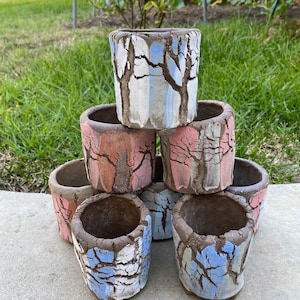 May include: A stack of seven cylindrical ceramic planters featuring a textured, cracked surface finish. Each planter displays a unique color palette including shades of blue, pink, and white, with dark brown clay accents forming tree-like patterns.