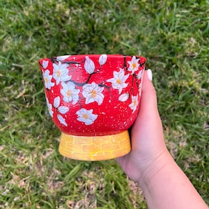 May include: A hand-painted red ceramic planter with white floral designs and a yellow base. The planter features a vibrant red background with white flowers and black branches. The base is a textured yellow brick pattern, perfect for indoor or outdoor use.