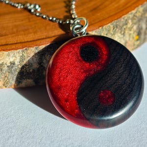 Könnte beinhalten: Eine Halskette mit einem Yin-und-Yang-Anhänger. Der Anhänger hat eine runde Form mit einem roten und schwarzen Design. Die Halskette ist silberfarben.