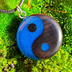 Puede incluir: Colgante redondo con diseño de Yin y Yang. El colgante presenta una combinación de colores azul y negro, con un punto negro en cada sección. Cuelga de un collar de cordón negro. Joyería.