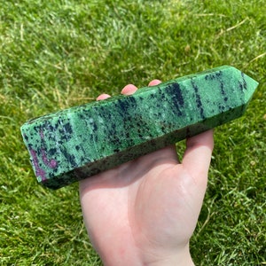 Może przedstawiać: Zielona i czarna różdżka z kryształu zoisytu z czerwonymi wtrąceniami. Polerowany kryształ ma prostokątny kształt ze spiczastym końcem. Różdżka jest trzymana na tle zielonej trawy.