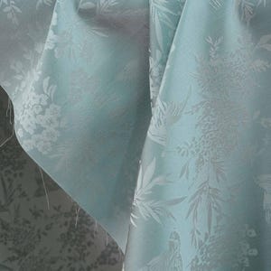 May include: Close-up of a folded, light blue fabric with a floral and bird pattern. The fabric has a subtle sheen and a detailed, embossed design. The pattern includes flowers, leaves, and birds.