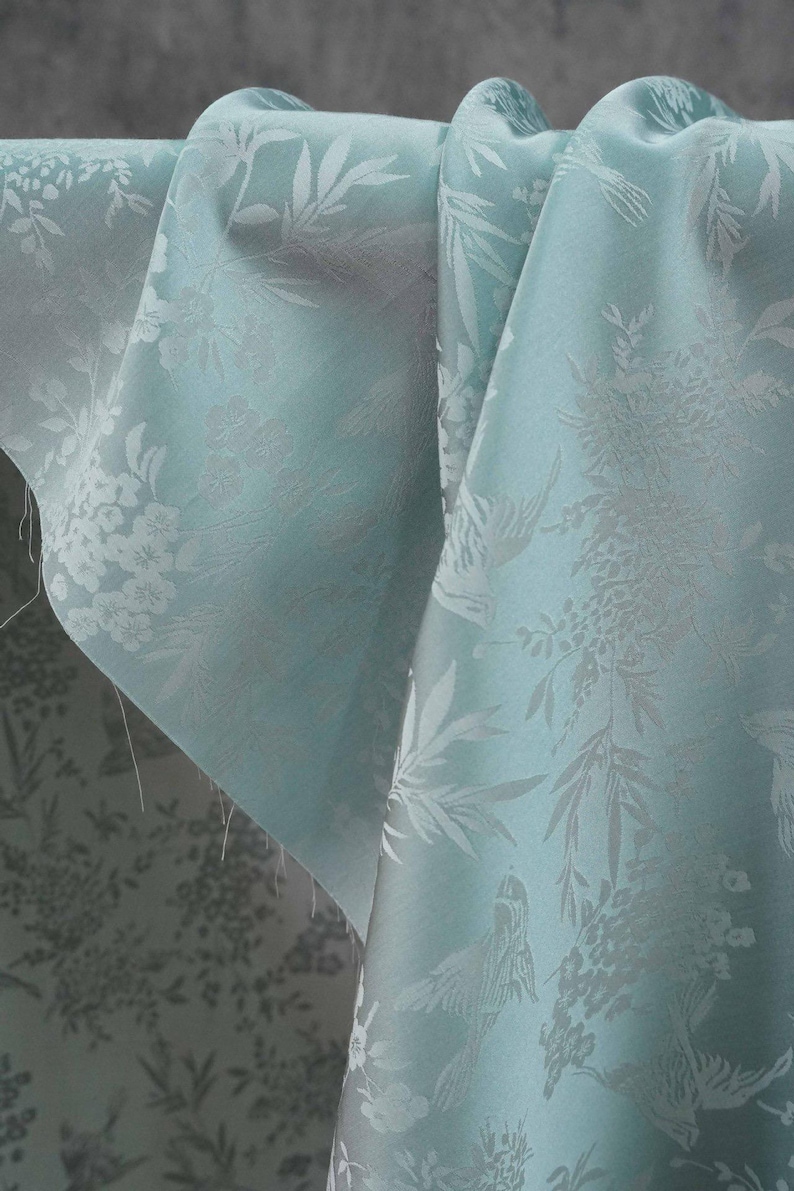 May include: Close-up of a folded, light blue fabric with a floral and bird pattern. The fabric has a subtle sheen and a detailed, embossed design. The pattern includes flowers, leaves, and birds.
