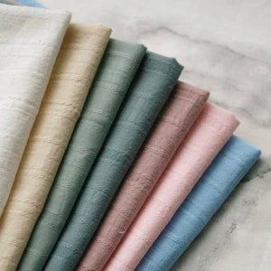 Puede incluir: Un conjunto de ocho muestras de tela en varios colores, incluyendo blanco, beige, azul claro, rosa y verde. La tela tiene un patrón texturizado sutil.