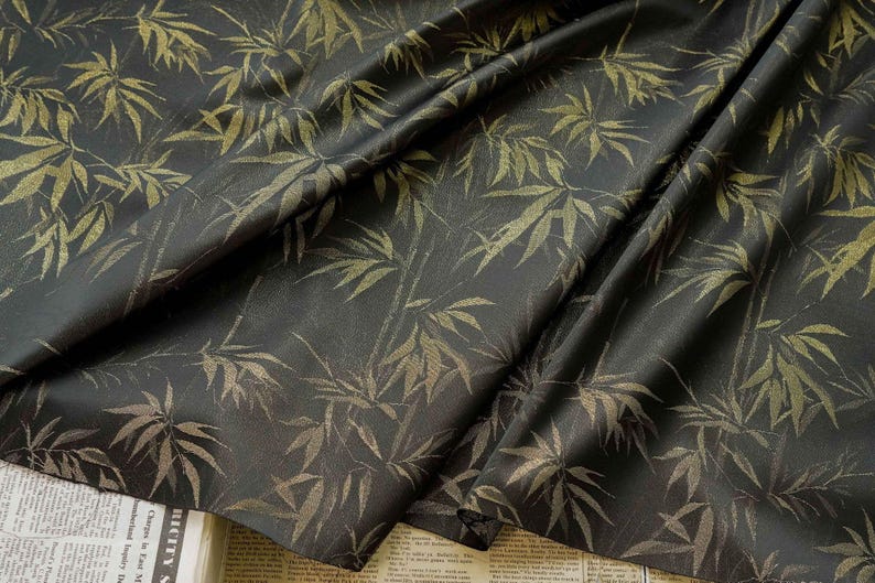 May include: Dark fabric featuring a gold bamboo leaf pattern. The leaves and stalks are in varying shades of gold, creating a textured effect against the dark background. The fabric is draped, showing folds and shadows.