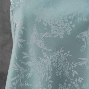 May include: A light blue fabric with a repeating pattern of birds, flowers, and foliage. The design is in a lighter shade of blue, creating a subtle contrast. The fabric has a soft sheen and a delicate appearance.