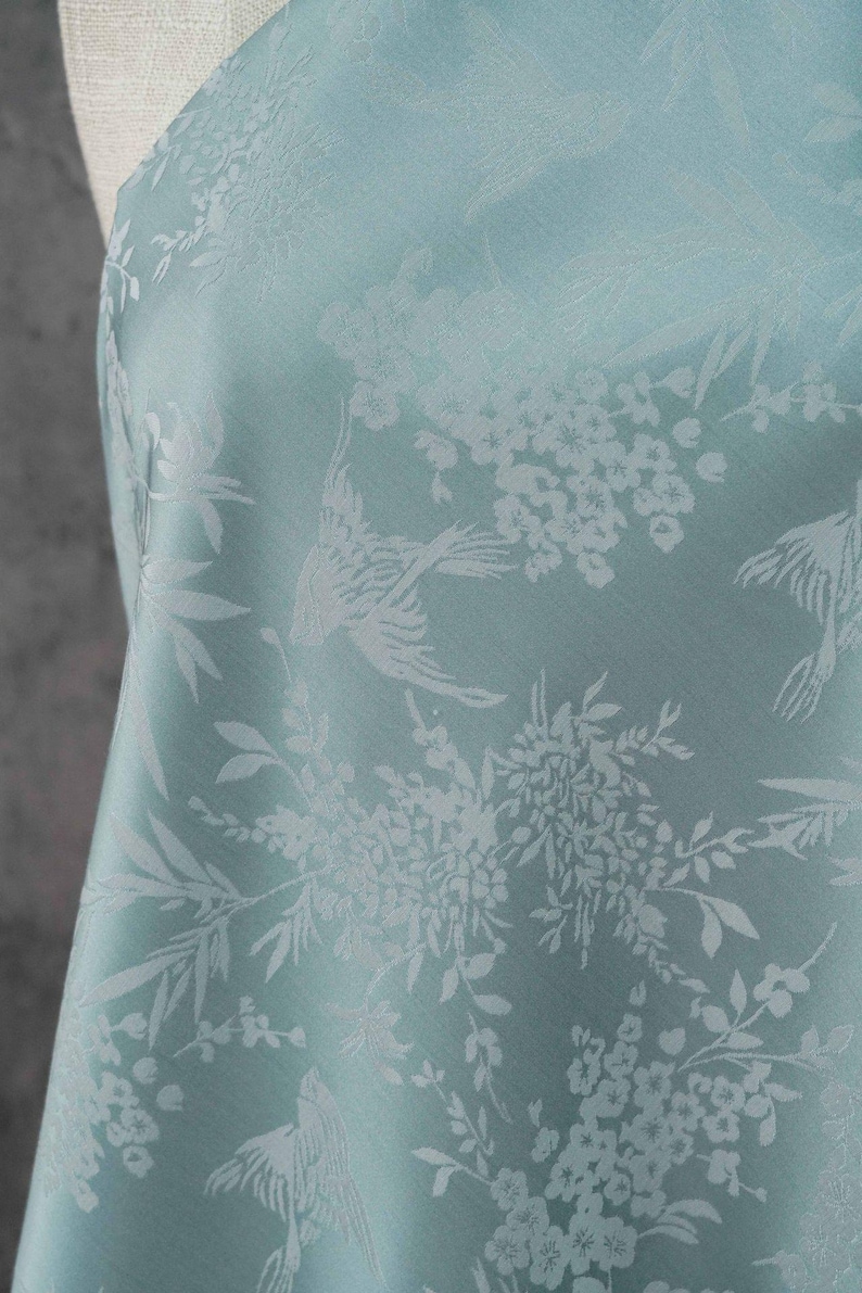 May include: A light blue fabric with a repeating pattern of birds, flowers, and foliage. The design is in a lighter shade of blue, creating a subtle contrast. The fabric has a soft sheen and a delicate appearance.