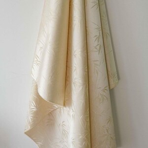 May include: Cream-coloured fabric with a subtle bamboo leaf pattern. The fabric is draped against a white wall, showcasing its texture and design. The fabric's folds create shadows, highlighting the pattern.