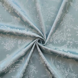May include: A close-up of a folded, light blue fabric with a floral and bird pattern in white. The fabric has a subtle sheen, and the design includes detailed depictions of flowers and birds, creating a delicate and elegant aesthetic.