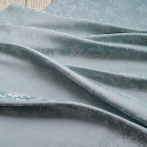 May include: A light blue fabric with a floral pattern. The fabric is draped, showing folds and shadows. The floral design is white and repeats across the material. The fabric is likely used for clothing or home decor.