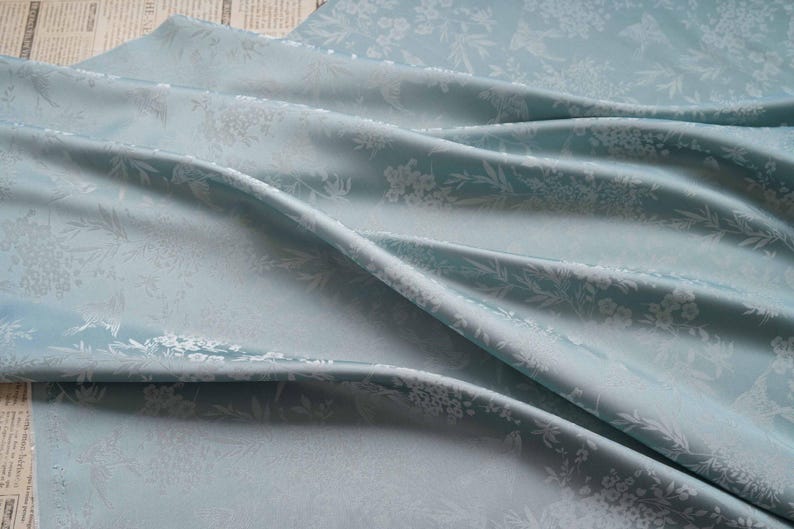 May include: A light blue fabric with a floral pattern. The fabric is draped, showing folds and shadows. The floral design is white and repeats across the material. The fabric is likely used for clothing or home decor.