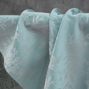 May include: Close-up of a light blue fabric with a floral and leaf pattern. The fabric is draped, showing the texture and design. The pattern is a lighter shade of blue, creating a subtle contrast.
