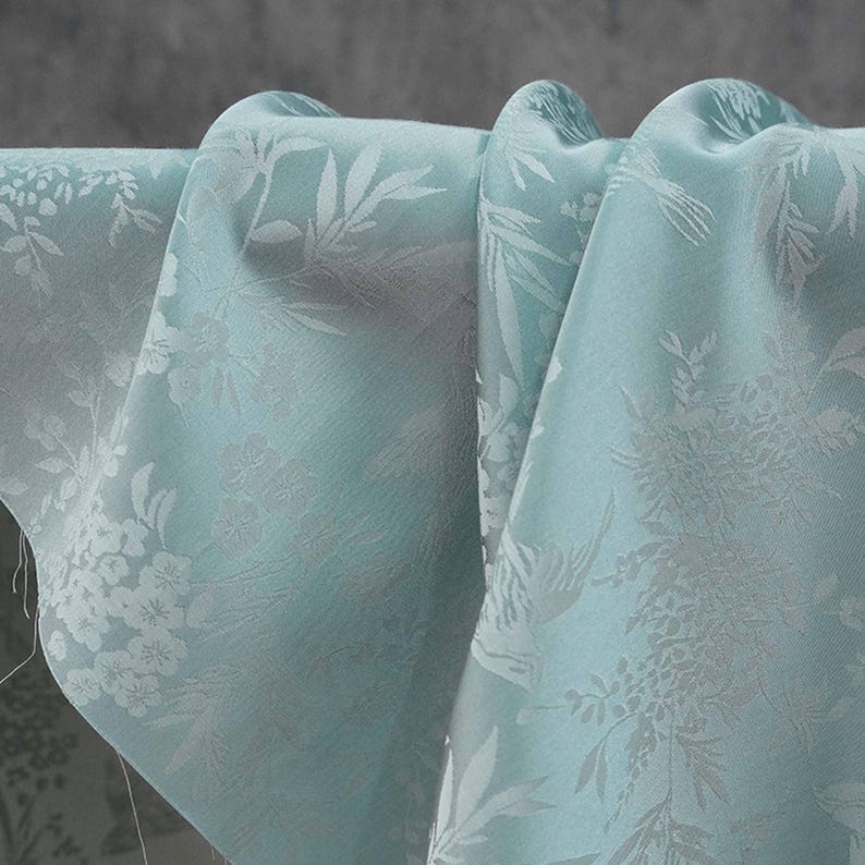 May include: Close-up of a light blue fabric with a floral and leaf pattern. The fabric is draped, showing the texture and design. The pattern is a lighter shade of blue, creating a subtle contrast.