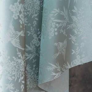 May include: A close-up of a fabric with a repeating pattern of birds and floral elements in white against a pale teal background. The fabric is folded, revealing the texture and design details. The overall aesthetic is delicate and elegant.