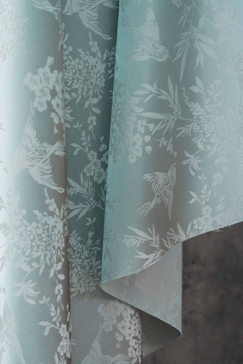 May include: A close-up of a fabric with a repeating pattern of birds and floral elements in white against a pale teal background. The fabric is folded, revealing the texture and design details. The overall aesthetic is delicate and elegant.