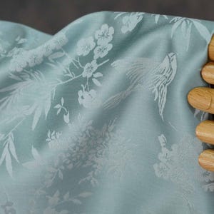 May include: Light blue fabric with a white floral and bird pattern. The fabric is draped, showing the intricate design of flowers, leaves, and birds. The fabric is held by a wooden hand.